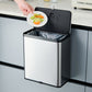 Hand dropping vegetable scraps from a plate into an open stainless steel trash can with a black lid