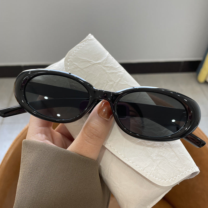 Black oval frame sunglasses with dark gray lenses held in a hand above an off-white glasses case