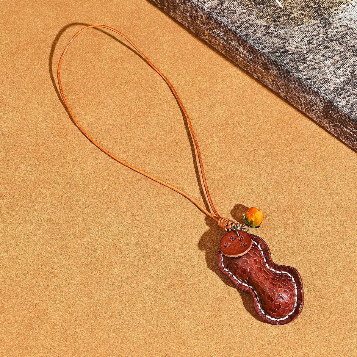 Dark brown peanut shaped leather pendant with a cord loop, orange glass bead and stamped tag on a tan background