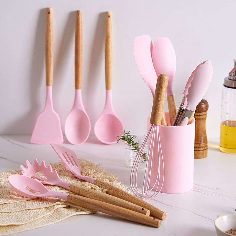 Pink utensil set styled on a kitchen counter with three tools hung on the wall and the rest in the holder
