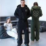 Man in a black zip hoodie and trousers beside a green hoodie back with a clover motif and a seated woman in gray
