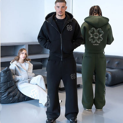 Man in a black zip hoodie and trousers beside a green hoodie back with a clover motif and a seated woman in gray