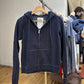 Navy zip-up hoodie with drawstring hood and raw edge seams on a clothes rack, with a size label inside the hood