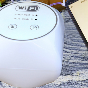 Automatic drip watering pump unit set beside a smartphone showing WiFi app control on a grey rug