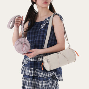 Woman in a plaid dress wearing a cream cylindrical nylon shoulder bag and holding a small lilac drawstring pouch