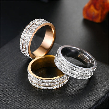 Three stainless steel band rings with rows of clear stones in rose gold, gold and silver tone finishes on a black surface