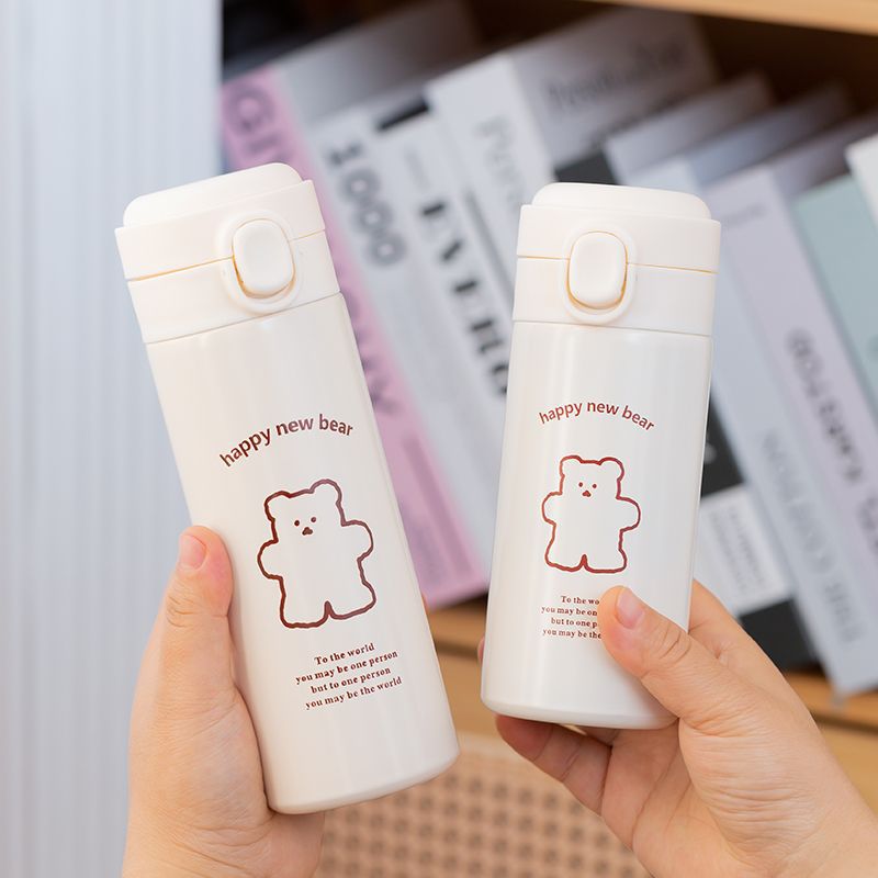 Two cream insulated bottles held in front of books, brown bear drawing and happy new bear text