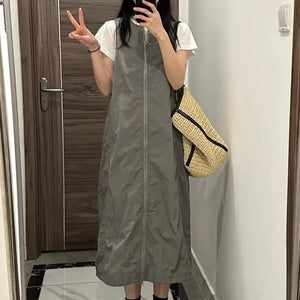 Woman in gray zip-front pinafore dress over white tee holding a straw bag