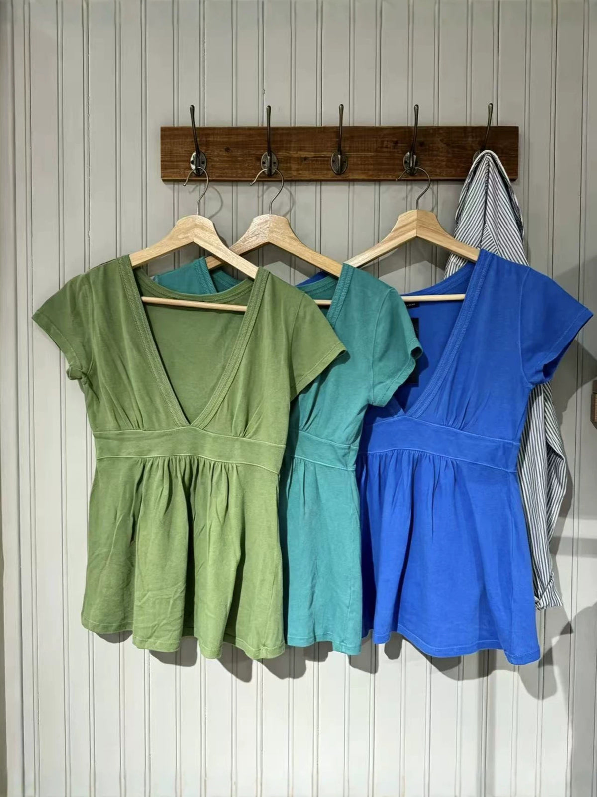 Three short-sleeve V-neck tops in green, teal and royal blue hanging on wall hooks