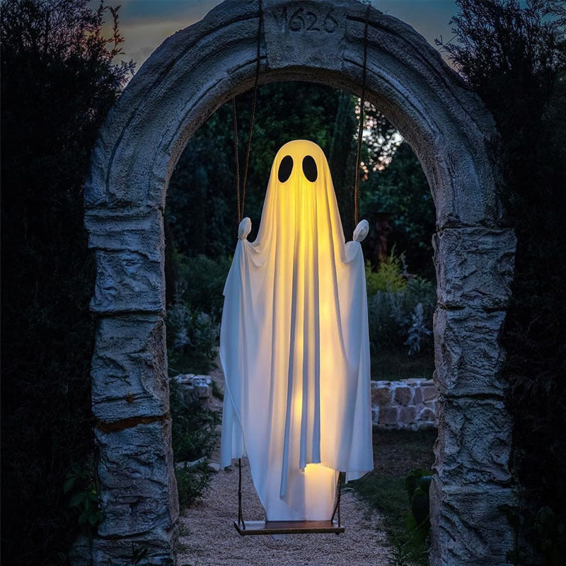 Lit white ghost on a rope swing under a stone arch in a garden at dusk, front view