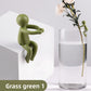 Grass green figure seated on a white block, with a small olive figure on a glass of water holding a flower stem
