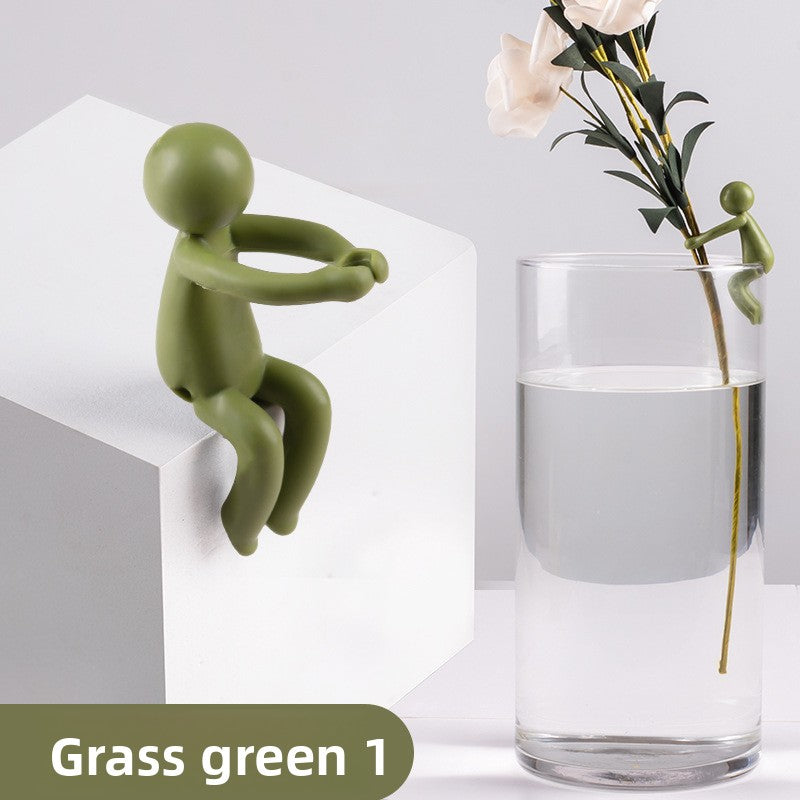 Grass green figure seated on a white block, with a small olive figure on a glass of water holding a flower stem