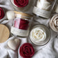 Rose shaped candles in glass jars with bamboo lids, a pink red rose and white roses on linen