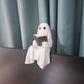 Resin ghost with a dark book sitting on a desk edge in front of a monitor and a teal curtain