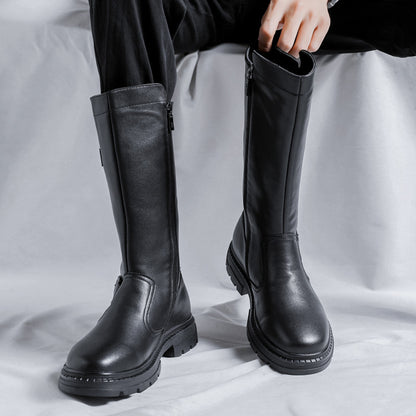 Pair of tall black boots with side zippers worn with black trousers, seated on a white drape