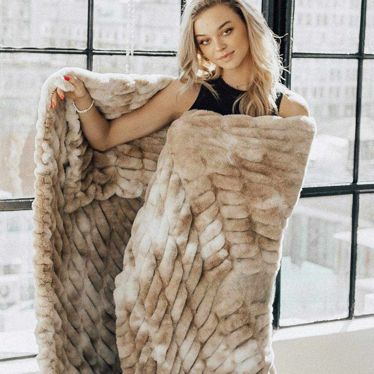 Woman by a large window holding up a ruched tie-dye brown plush blanket draped over her shoulder