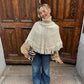 Smiling woman in a cream fringed knit poncho top over a beige blouse and wide jeans in front of wooden doors