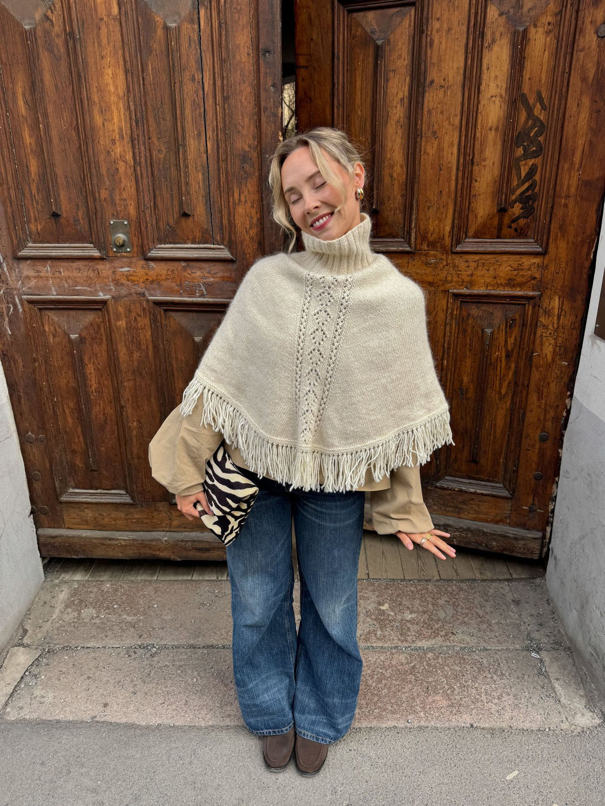 Smiling woman in a cream fringed knit poncho top over a beige blouse and wide jeans in front of wooden doors