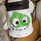 White party bucket bag with a stitched green monster face and a black carry handle