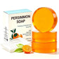 Three stacked amber persimmon soap bars beside a Persimmon Soap box, a white mesh net and a halved persimmon