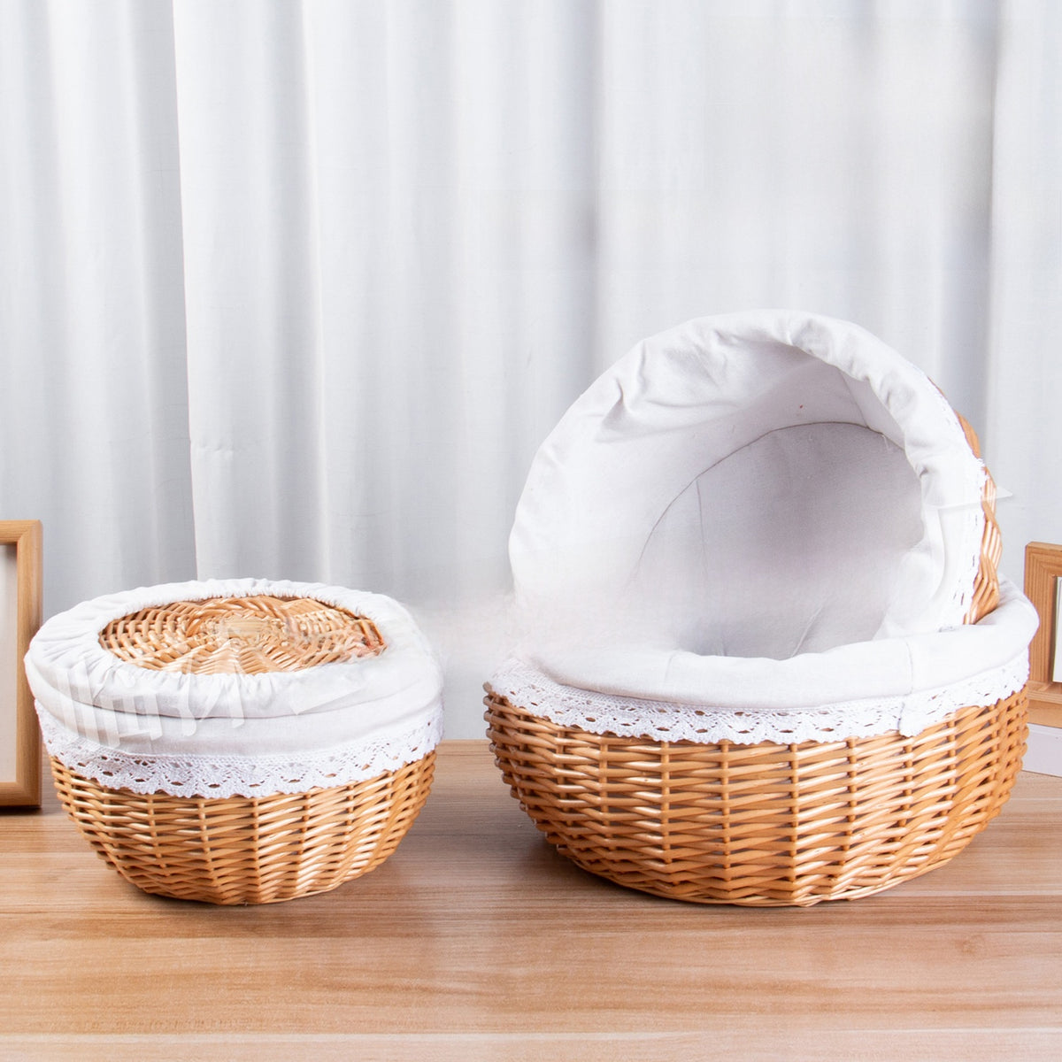 Two round wicker baskets with white fabric lining and lace trim, one closed with its lid, on a wooden table