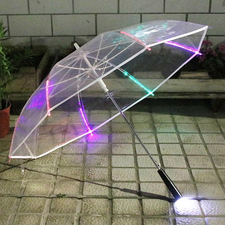 Clear umbrella with a white canopy edge and pink, green and red lit rib segments on a tiled courtyard at night