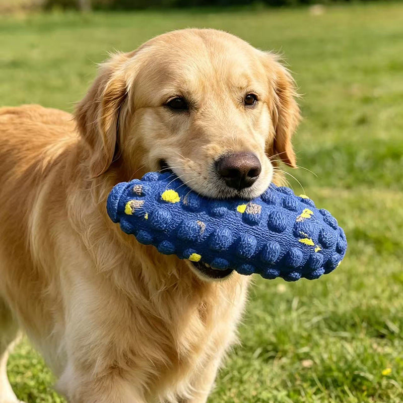 Golden retriever carrying a blue bumpy corn cob shaped dog toy with small yellow flecks across a green lawn