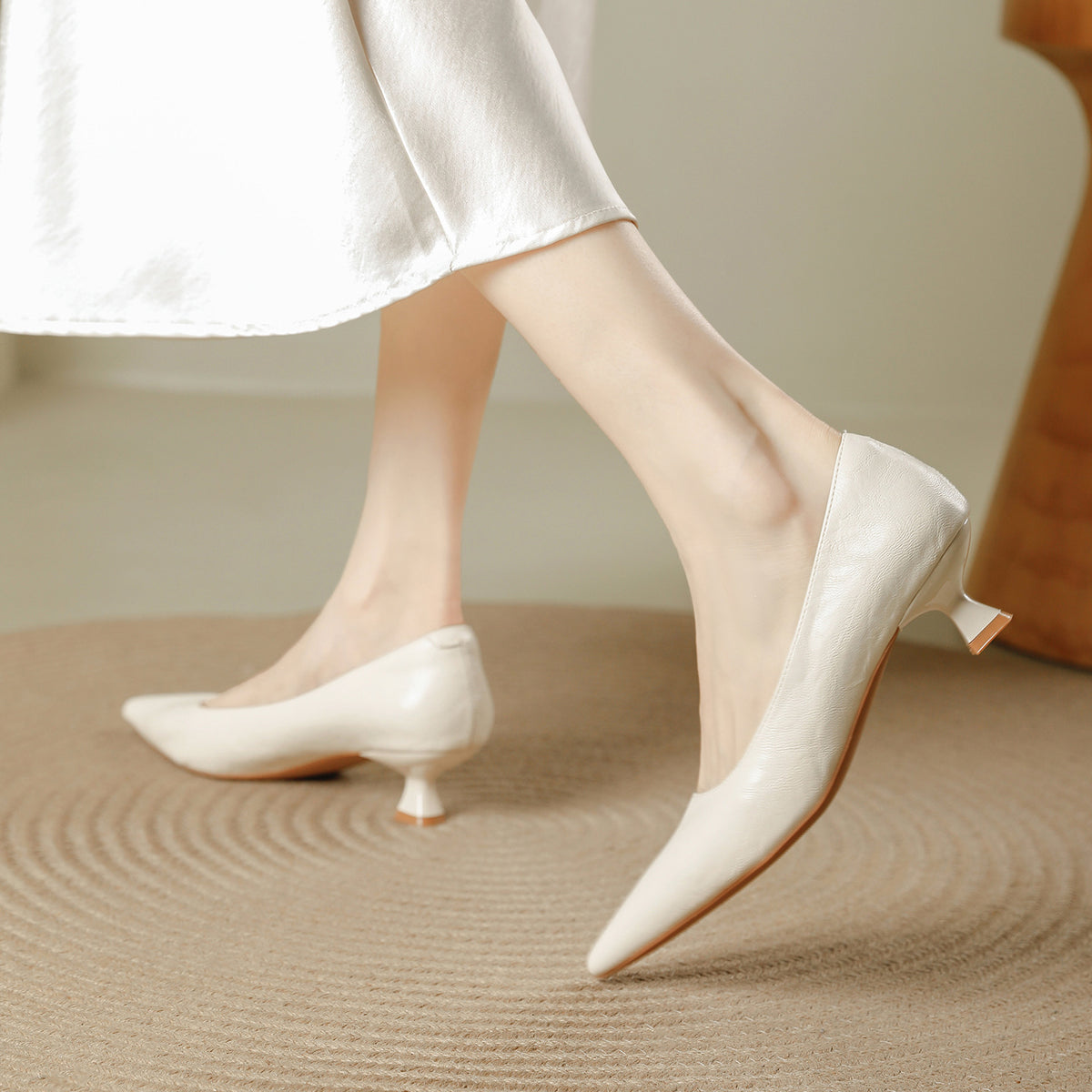 Cream pointed toe pumps with a low flared heel worn with a white skirt on a woven rug