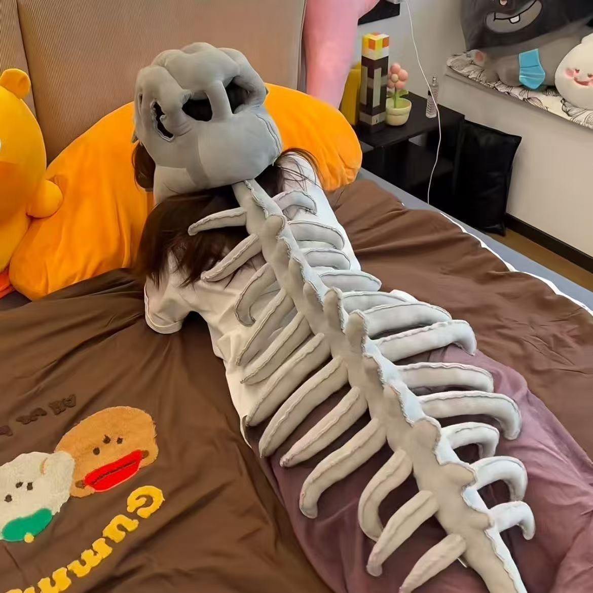 The grey dinosaur skeleton plush hood laid flat, showing the skull and segmented spine