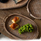 Oval woven rattan tray holding two croissants and green grapes, studded wooden rim and arched handle