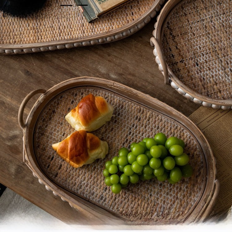 Oval woven rattan tray holding two croissants and green grapes, studded wooden rim and arched handle