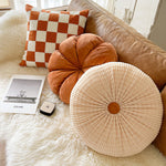 Checkerboard, pumpkin and round pleated plush pillows in orange and beige on a brown leather sofa with a white throw