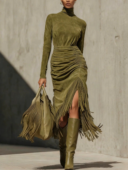Woman in an olive green mock neck dress with ruched skirt and fringe slit, olive boots and bag, concrete wall