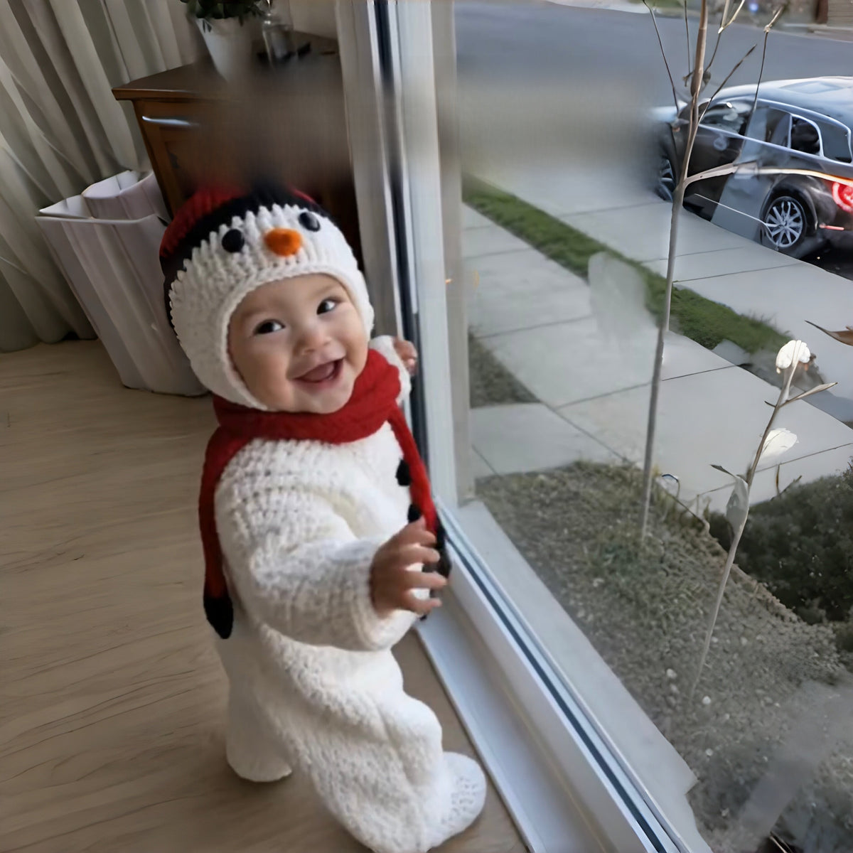 Toddler in a white knitted snowman hat and red scarf standing by a window, snowman style not offered