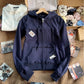 Navy zip-up hooded sweatshirt on a wooden hanger with raw edge seams, a pocket and a drawstring hood