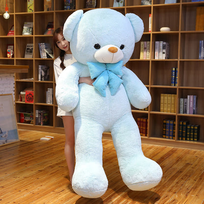 Light blue plush teddy bear with a blue striped bow tie standing next to a smiling girl in front of a bookshelf