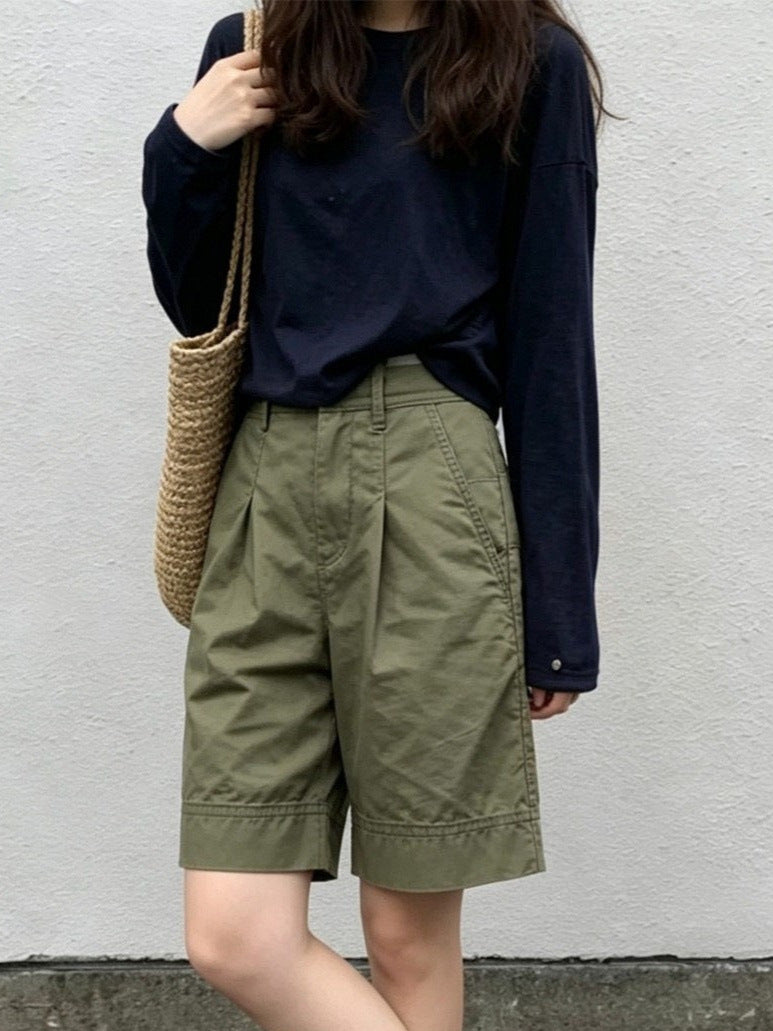 Front view of woman in olive green high-waisted Bermuda shorts with navy tee and woven bag