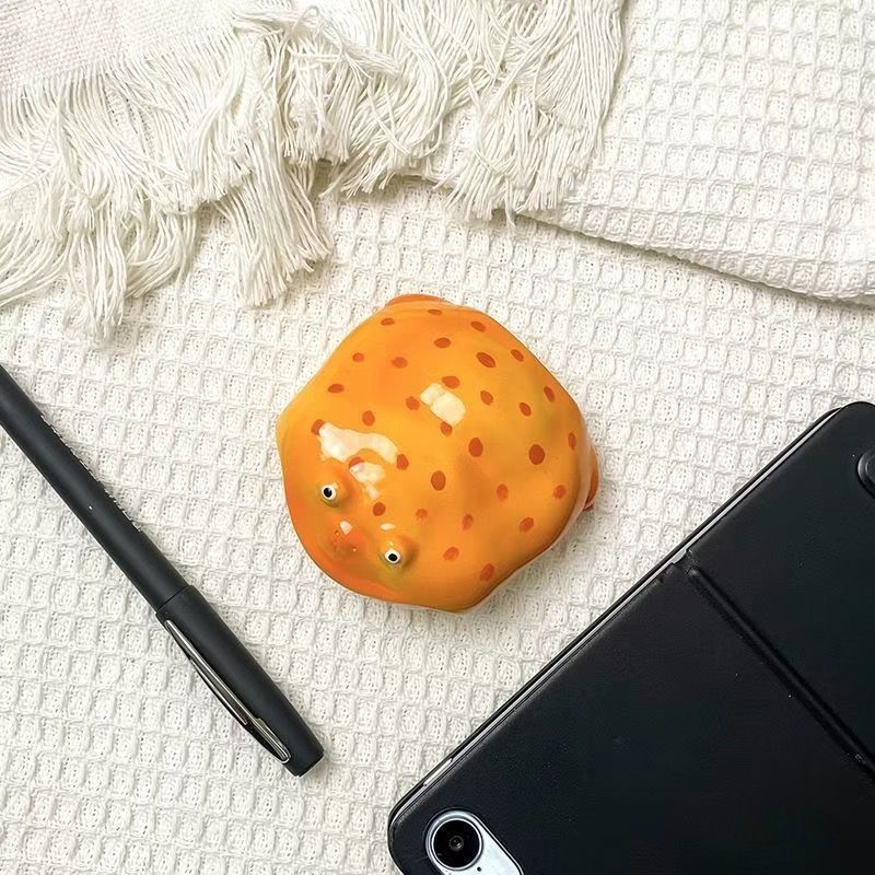 Plain orange resin frog earbud case seen from above on white cloth beside a pen and a tablet
