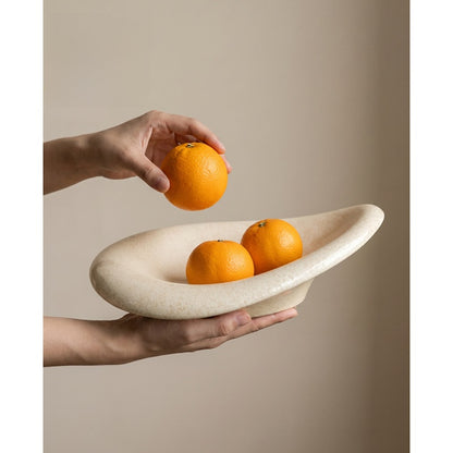 Hands placing an orange into a glossy beige ceramic oval fruit bowl holding two oranges