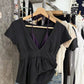 Black short-sleeve deep V-neck top with gathered hem on a hanger over a rail of clothes