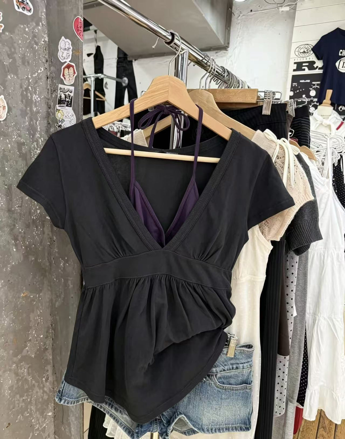 Black short-sleeve deep V-neck top with gathered hem on a hanger over a rail of clothes