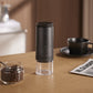 Black electric coffee grinder with a top display disc beside a jar of coffee beans, a black cup and a newspaper