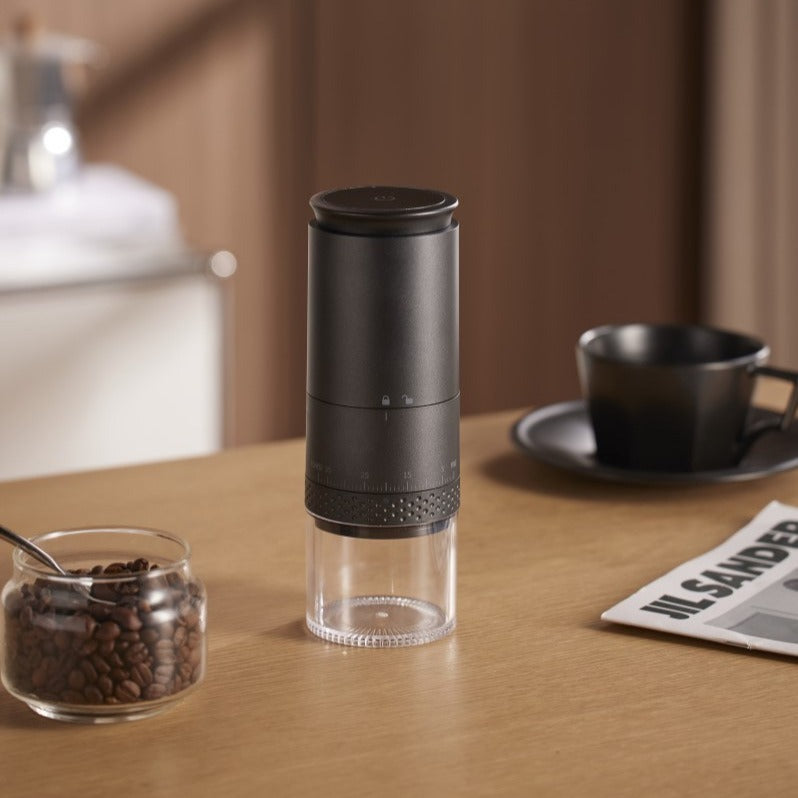 Black electric coffee grinder with a top display disc beside a jar of coffee beans, a black cup and a newspaper