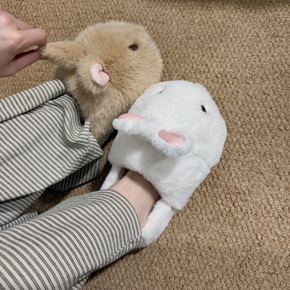 One brown and one white bunny plush slipper with pink ears worn with striped trousers on a woven carpet, view 1 of 3