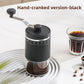 Black hand-cranked coffee grinder with crank arm, fine to coarse scale and clear grounds chamber beside a coffee glass