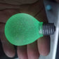 Glow option of the squeeze toy, bulb shape lit flat green in the dark against a hand