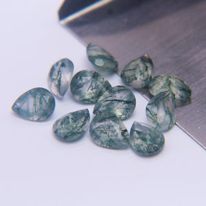Eleven faceted moss agate pears scattered beside a steel bench blade on white