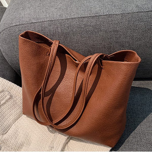 Tan brown PU tote bag with two long straps and a soft pebbled surface resting on a gray sofa