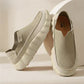 Khaki perforated slip-on loafers with a cream scalloped sole and a tan insole, one standing against a round beige backdrop