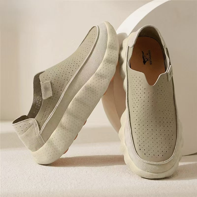 Khaki perforated slip-on loafers with a cream scalloped sole and a tan insole, one standing against a round beige backdrop